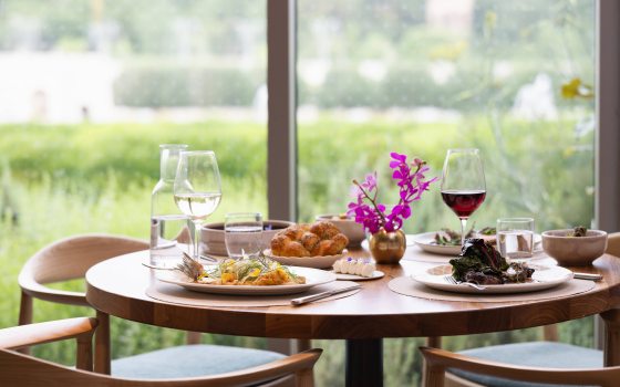 A round dining table set with food, drink, and flowers, against a backdrop of bright windows with a view of a fountain garden.