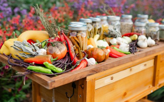 Closeup of wooden cart laden with fresh vegetables and filled mason jars, against a garden backdrop of red and purple blossoms.