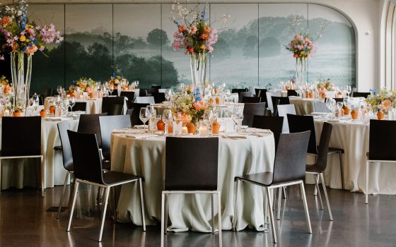 Seven tables are set for guests to dine at with linens, plates, and tall floral arrangements in a room with shiny black floors and a green mural on the back wall. 
