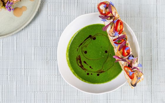 An overhead shot of a bowl of bright green soup with a dark oil drizzle. A long baguette slice sits on the edge, decorated with mussels, red chili rings, and delicate purple and blue floral garnishes. A corner of another plate with a small appetizer is visible in the top left.