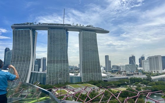 A wide view taken from the top of the Supertree Grove in Singapore, looking out toward the iconic Marina Bay Sands hotel. The three skyscrapers with their connecting surfboard-shaped rooftop garden dominate the skyline, set against a bright, hazy blue sky with light clouds. In the foreground, the purple structural ribs of a Supertree and solar panels are visible.