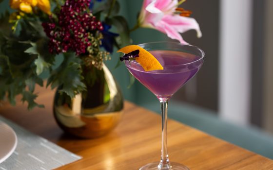 A long-stem cocktail glass holds a purple drink garnished with a flower and orange peel, with a gold-colored vase of summer flowers in the left background.
