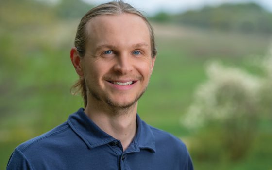 A man with blonde hair wearing a navy shirt smiles in front of a green and grassy meadow. 