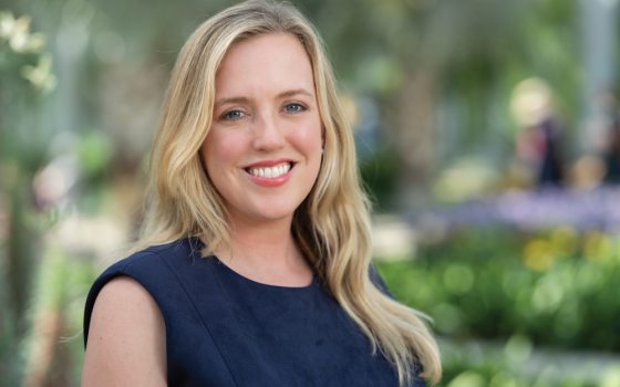 A woman with blonde hair wearing a navy shirt smiles in front of a backdrop of lush green plants 