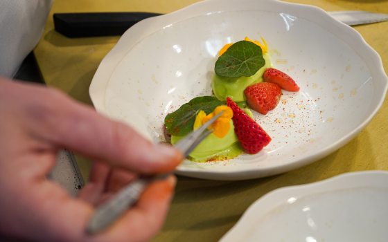 An over-the-shoulder close-up of a chef using kitchen tweezers to place a delicate orange edible flower petal onto a plated dish consisting of vibrant green sorbet, sliced fresh strawberries, and green nasturtium leaves in a wide white ceramic bowl.