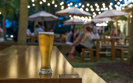 A glass of beer sits on the edge of a picnic table, against a background of guests at tables under party lights and table umbrellas.