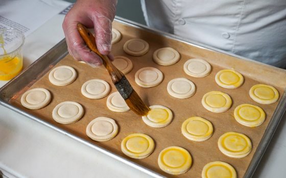 A baker wearing plastic gloves uses a pastry brush to apply a golden egg wash onto rows of small, circular, unbaked pastries arranged on a parchment-lined baking sheet.