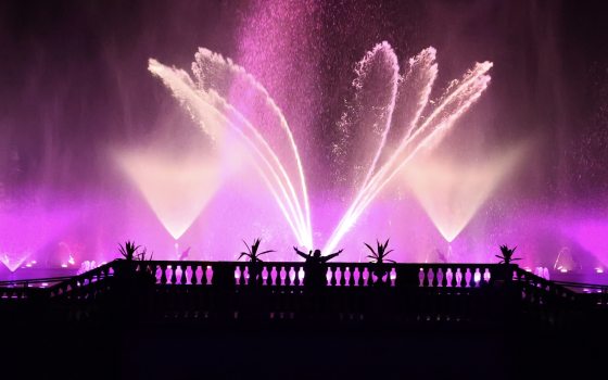 Silhouette of a person standing with arms raised on a balcony, watching a grand, fan-shaped nighttime fountain display illuminated in a deep magenta hue.