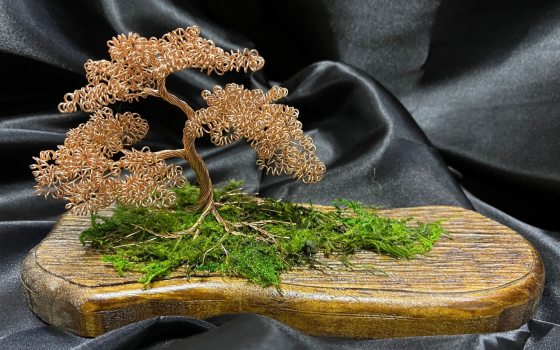 A wire bonsai sculpture on a wooden board sitting against a black fabric backdrop.