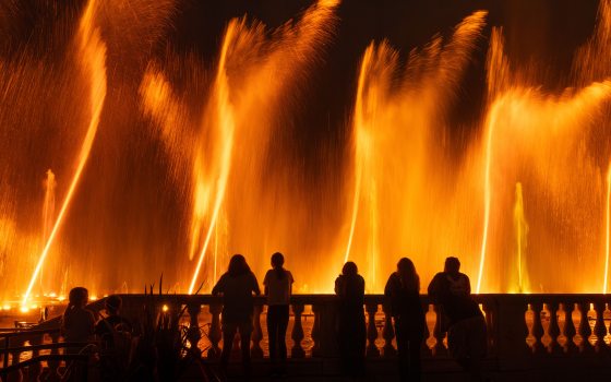 Guests in black silhouette stand at a rail watching orange fountains rising like flames.