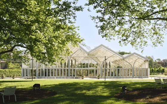 Large trees shade a green lawn in front of a large glass house with peaked arches.