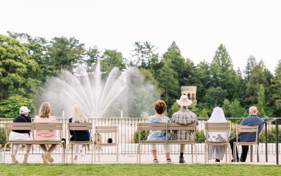 People seated outdoors in tan chairs, watching fountains in front of them. 