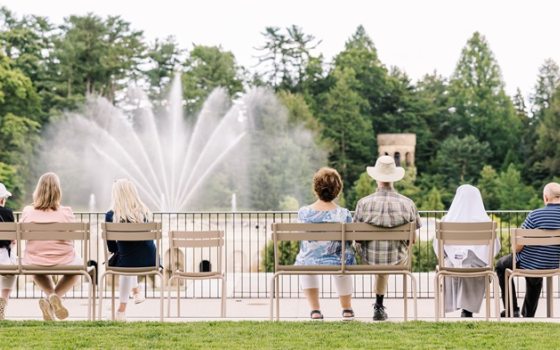 People seated outdoors in tan chairs, watching fountains in front of them. 
