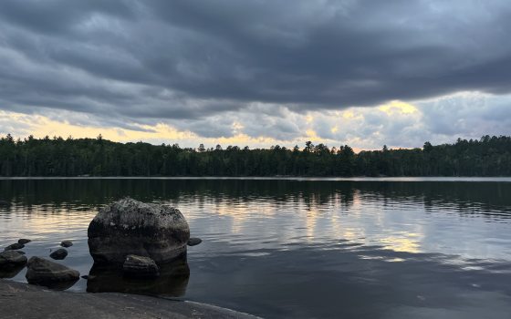A tranquil lake reflects a dramatic sunset through dark, heavy storm clouds hovering over a distant, densely forested shoreline, with a large rounded boulder resting in the water near the rocky foreground.