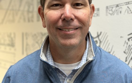 A man with black hair wearing a light blue shirt and a nametag smiles in front of a white wall with grey sketches on it. 