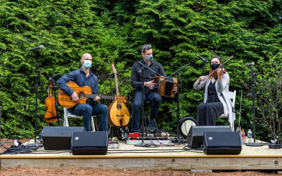 a group of three musicians performing on a small stage with trees in the foreground