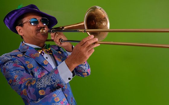 A musician in a colorful floral jacket, sequined bowtie, purple hat, and tinted glasses plays the trombone