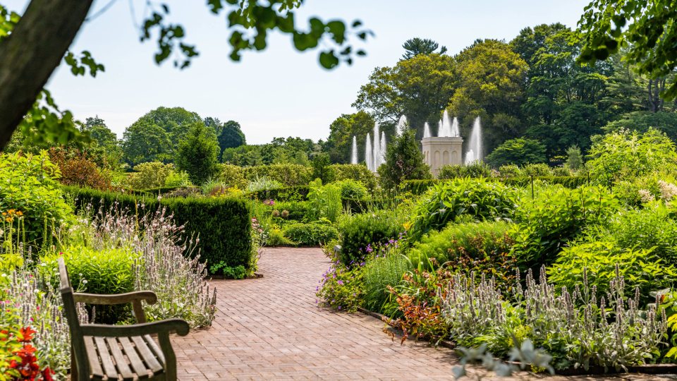 A brick walkway with a wooden bench winds through green garden beds with fountains in the distance