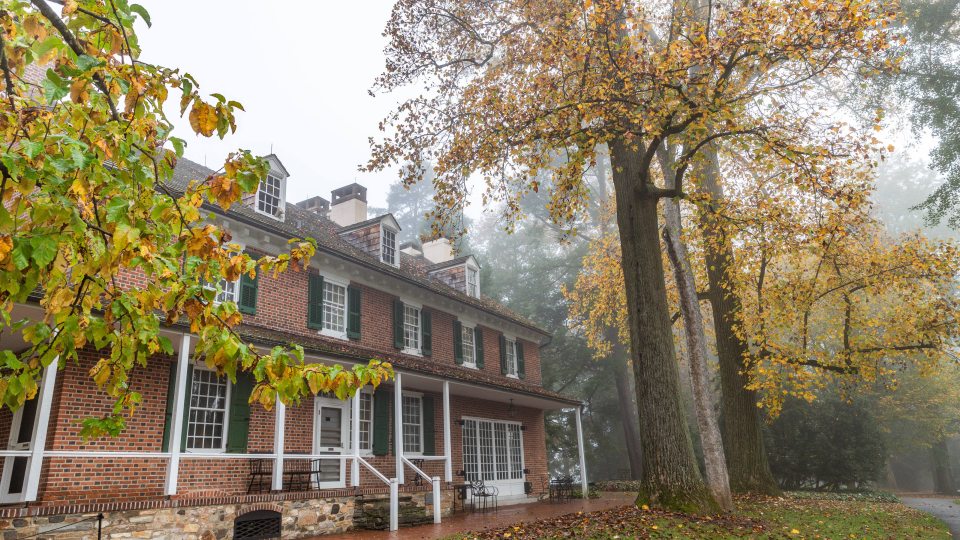 A stone and brick house is seen surrounded by autumn trees of yellow and orange and a morning fog