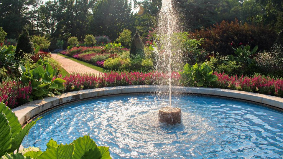 Sun shines on a circular fountain with a brick pathway in the background leading through green garden beds