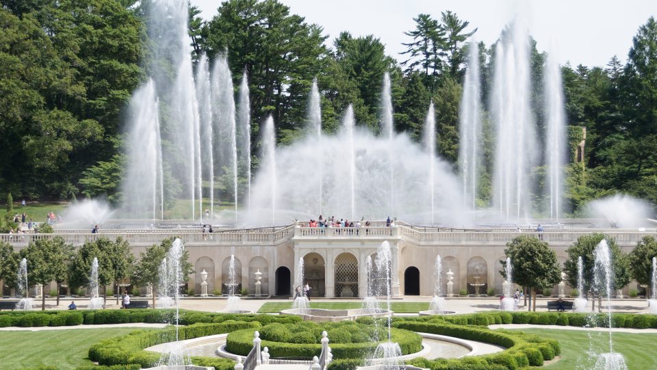 Fountains shoot water high into the air during the day over carved white stone structure and green boxwoods