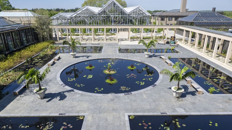 Broad view of one central round waterlily pool, surrounded by stone walkways, rectangular pools, and conservatory structures.