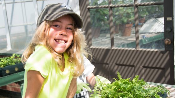 Child with long blonde hair, lemon-lime t-shirt, and a baseball cap smiles broadly in front of a potting bench with green plants