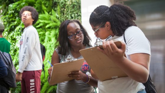 Students with clipboards work together in front of a wall of green plants
