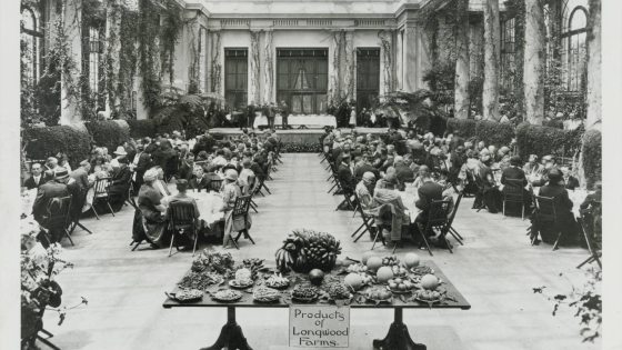 Black and white historical image of guests seated at long dining tables in a lush indoor garden setting, with a table in front filled with produce, with a sign that reads "Products of Longwood Farms"