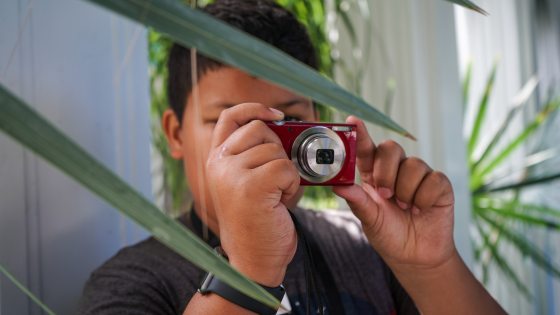 a young person looks through a small red camera, facing palm fronds