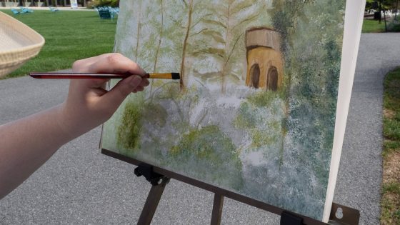A hand holding a paintbrush works on a canvas of a watercolor painting of trees and a carillon tower, with that landscape in the background, including an arched stone wall of fountains not included in the painting.