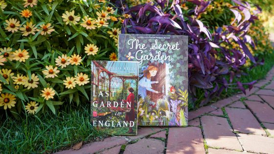 Two books (The Last Garden in England, and The Secret Garden) are propped up against a bed of yellow flowers and purple foliage, along a curved brick path.