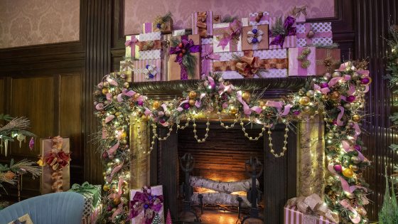 An ornate fireplace, with birch logs aglow, is decked out with gold ball garland, ornaments in pink and gold, and festive presents in pink and gold tones piled high on the mantel, and all around the hearth.