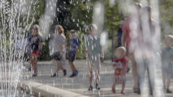 People of all ages stroll through a fountain garden, as seen through the spray of the fountains.