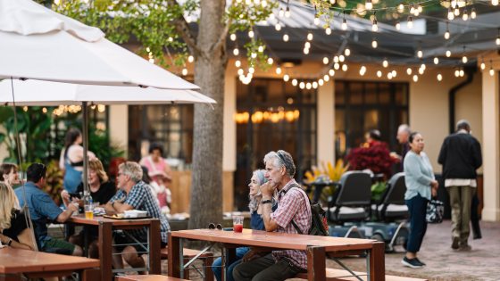 People eat and drink at umbrella-covered tables under strings of lights in an outdoor setting.