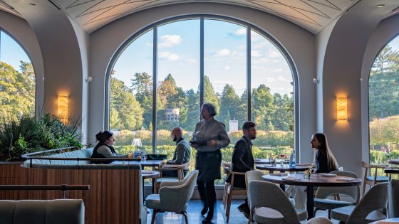 View of a diners seated in a restaurant in front of a large arched window that looks out onto fountains and colorful autumn trees in the distance. 