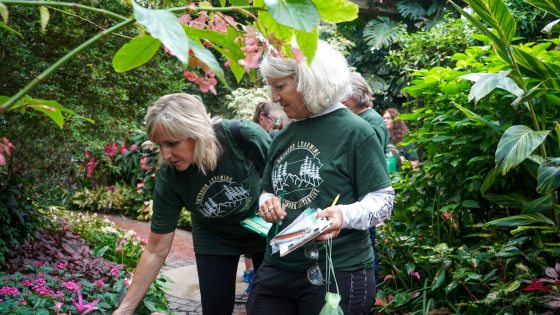 Two educators in green t-shirts that say "Outdoor Learning" examine plants along an indoor garden path.