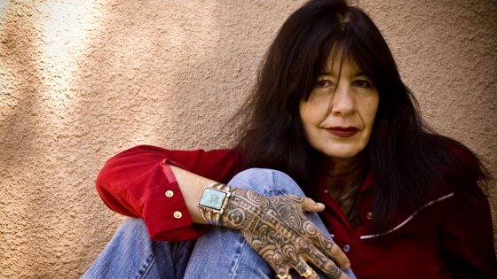 Women in red shirt and jeans sits facing the camera and displaying an intricate tattoo on her hand and a turquoise cuff bracelet 