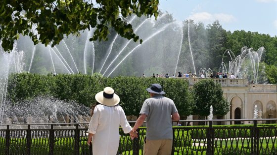 A couple wearing summer garden hats hold hands and watch a daytime fountain show from behind an ornamental metal fence.