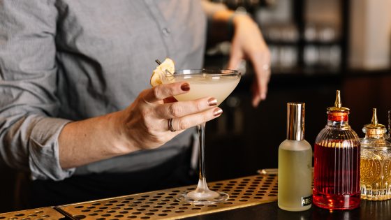A person's hands behind a bar holding a cocktail glass preparing to hand it to someone. 