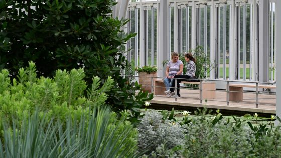 Two people sit on a bench in a conservatory amid lush green plants and soaring arches.