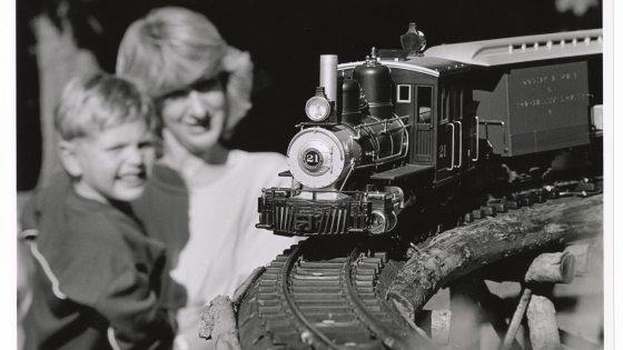 A black and white photograph of a toy or model steam train engine and car running on a curved track in the foreground. In the soft-focus background, an adult woman and a young boy are smiling as they watch the train.