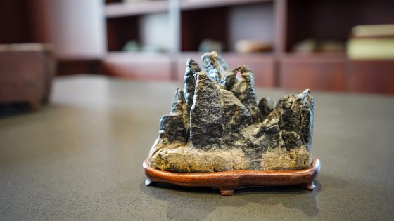  The alt text for this image should be:  A close-up, low-angle shot of a small, dark, craggy rock formation carving, resembling miniature mountains or peaks. It sits on a smooth, dark wooden stand with curved feet. The object is centered on a grey, textured table, with a blurred background of dark wooden shelving or cabinetry.