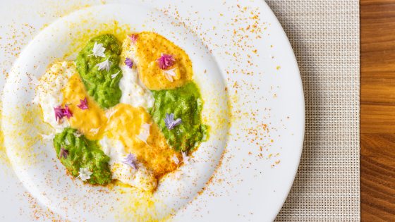 Close-up, overhead view of a plated dish, likely risotto, with alternating dollops of creamy white, vibrant green, and yellow puree or sauce, garnished with edible flowers and surrounded by spice dust.