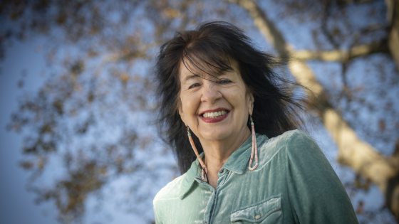 Outdoor portrait of poet Joy Harjo smiling at the camera, wearing a light teal velvet collared shirt and long beaded earrings, with a large tree and clear blue sky out of focus behind her.