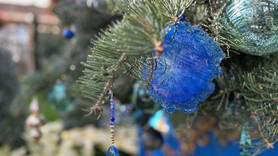 A close-up shot of a Christmas tree branch adorned with blue and turquoise ornaments. A striking, fuzzy, royal blue floral or star-shaped ornament is visible, along with a hanging decoration featuring a dangling, tear-drop-shaped clear blue crystal bead. White poinsettias are visible and slightly blurred in the lower left background