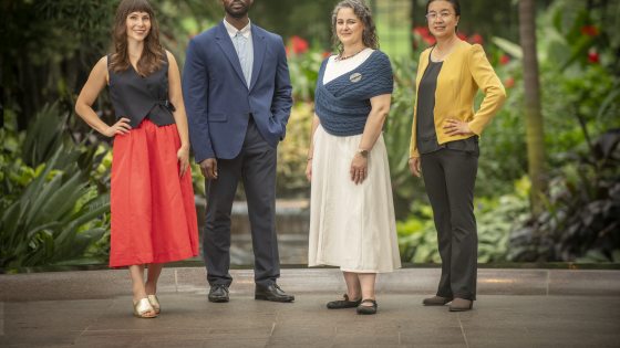 A full-body group portrait of four Longwood Fellows standing side-by-side in formal business-casual attire against a lush green background.