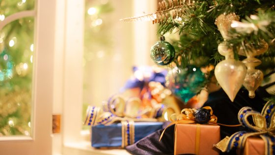 Closeup of ornate packages beneath a decorated Christmas tree, with guests reflected in a green ball ornament.