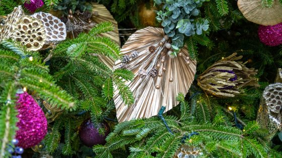 A close-up of a richly decorated Christmas tree showing a variety of ornaments, including large, pleated, metallic gold fan-shaped decorations, natural elements like pine sprigs and small pinecones, and bright, glittery ornaments in shades of magenta, purple, and gold.