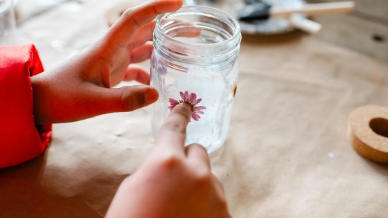 A close-up shot of hands carefully pressing a small, pink dried flower onto the side of a glass jar, showing the intricate detail of the craft project.